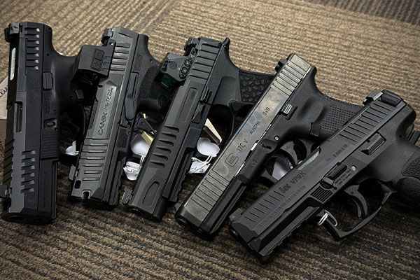 Five black handguns are arranged side by side on a textured, brown and beige carpeted surface.